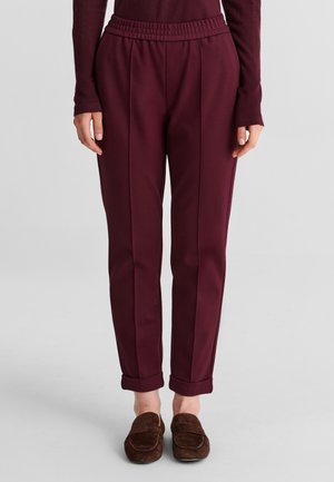 Personne portant un pantalon bordeaux fuselé avec coutures avant et revers roulés, associé à des mocassins en daim marron, debout devant un fond uni.