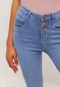 Jean taille haute bleu clair avec trois boutons à l'avant, poches latérales et texture en denim lisse.