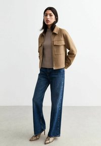 Giacca in suede marrone con colletto e due tasche sul petto, abbinata a un maglione beige a coste e jeans in denim a gamba larga. Tacchi in stampa serpente.