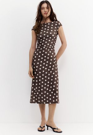 Femme en robe midi marron à pois blancs, manches courtes, taille cintrée, et sandales à talons noires, debout contre un fond blanc uni.