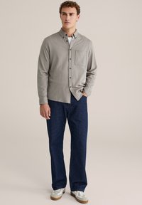 Lichtgrijs overhemd van zachte stof met een borstzak, gedragen met donkerblauwe spijkerbroek en grijs-witte sneakers. Recht gesneden pasvorm.