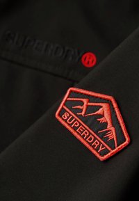 Zwarte stof met een rood geborduurd Superdry-patch met een hoekig ontwerp en gestileerde dierenillustraties. Subtiel logo in het zwart.