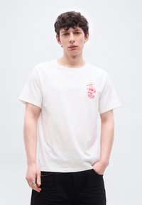 Νεαρός άνδρας με σγουρά μαλλιά που φοράει λευκό μπλουζάκι με κόκκινο γραφικό σχέδιο και κείμενο «Κρατώντας τα ψωμάκια ζεστά», με το χέρι στην τσέπη μαύρου παντελονιού.