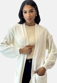 Kremowy kardigan o otwartym splocie z długimi rękawami, żebrowanymi akcentami i przednimi kieszeniami, noszony na beżowej, żebrowanej bluzce.