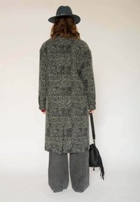 Long manteau à carreaux noirs et blancs avec une silhouette oversize, associé à un pantalon gris ample et un sac à main noir. Un chapeau gris complète le look.