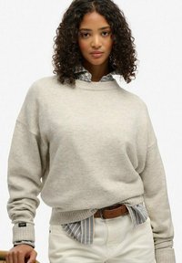 Suéter oversize gris claro con dobladillo acanalado, combinado con una camisa de rayas azul y blanca. La modelo lleva un cinturón marrón y pantalones crema.