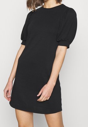 Kobieta w krótkiej czarnej sukience z bufkami na krótkich rękawach i okrągłym dekoltem, stojąca na tle jednolitego jasnego tła.