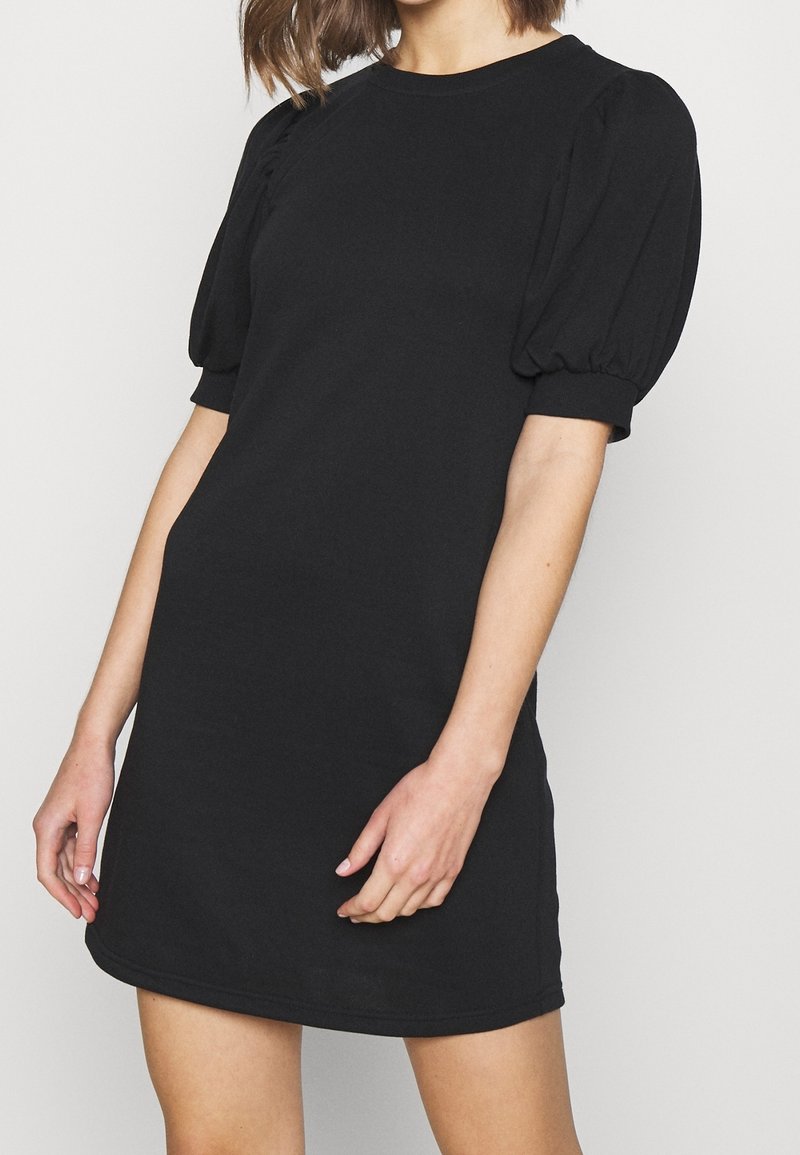 Femme portant une courte robe noire avec des manches courtes bouffantes et un col rond, debout devant un fond clair uni.