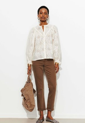 Femme debout contre un mur blanc, portant un chemisier blanc à œillets, un pantalon marron, des ballerines à imprimé léopard, tenant une veste marron pliée dans la main gauche.