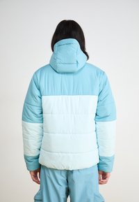 Vadderad jacka i ljusblå och blekblå nyanser med en huva, med horisontella quiltade sektioner och en slät yta.