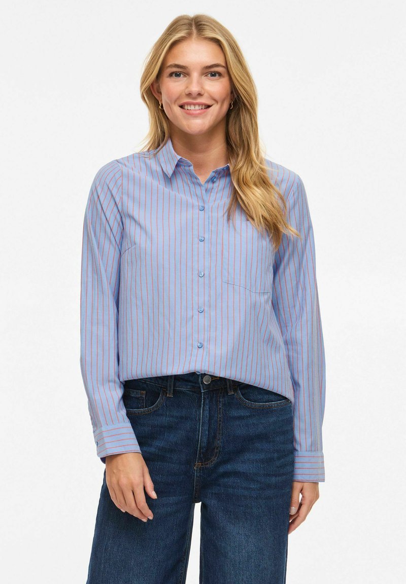 Jeune femme souriante, portant une chemise boutonnée bleu clair à fines rayures rouges et un jean bleu foncé, debout devant un fond blanc uni.