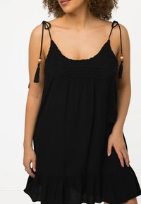 Vestido negro sin mangas con un corpiño texturizado, dobladillo con volantes, y lazos ajustables con borlas y cuentas de madera en las tirantes.