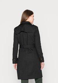 Cappotto lungo nero con vita a cintura, spalline sulle spalle e polsini abbottonati. La vista posteriore mostra una silhouette liscia e sartoriale con dettagli di cucitura.