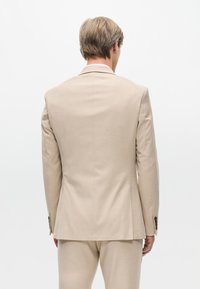 Beige maßgeschneiderte Anzugjacke mit glatter Textur, einer Mitteventilation hinten und Knopfdetails an den Ärmeln. Schlichtes Design, minimalistische Details.