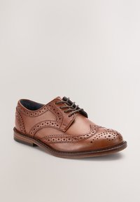 Bruine leren brogues met decoratieve perforaties en donkerbruine veters, op een lichte achtergrond.