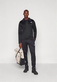 Μαύρο πουλόβερ με φερμουάρ και παντελόνι jogger με λογότυπο, υλικό με υφή πλεκτού. Το μοντέλο κρατάει ένα μπεζ σακίδιο πλάτης με μαύρες λεπτομέρειες. Μαύρες μπότες ολοκληρώνουν την εμφάνιση.