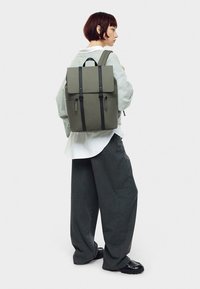 Sac à dos gris de forme structurée, avec des bretelles et des accessoires noirs, en matériau lisse et fermé par un rabat. Modèle portant un pantalon gris à coupe décontractée.