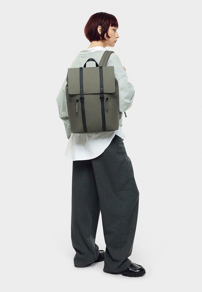 Sac à dos gris de forme structurée, avec des bretelles et des accessoires noirs, en matériau lisse et fermé par un rabat. Modèle portant un pantalon gris à coupe décontractée.