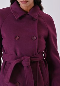 Donna che indossa un cappotto di lana viola scuro a doppio petto con cintura annodata e ampio colletto, mostrata dalle spalle alla vita.