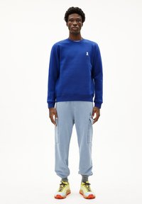 Blauwe sweatshirt met wit lintlogo, gecombineerd met lichtblauwe cargobroek. Heldere sneakers met gele, oranje en zwarte accenten.