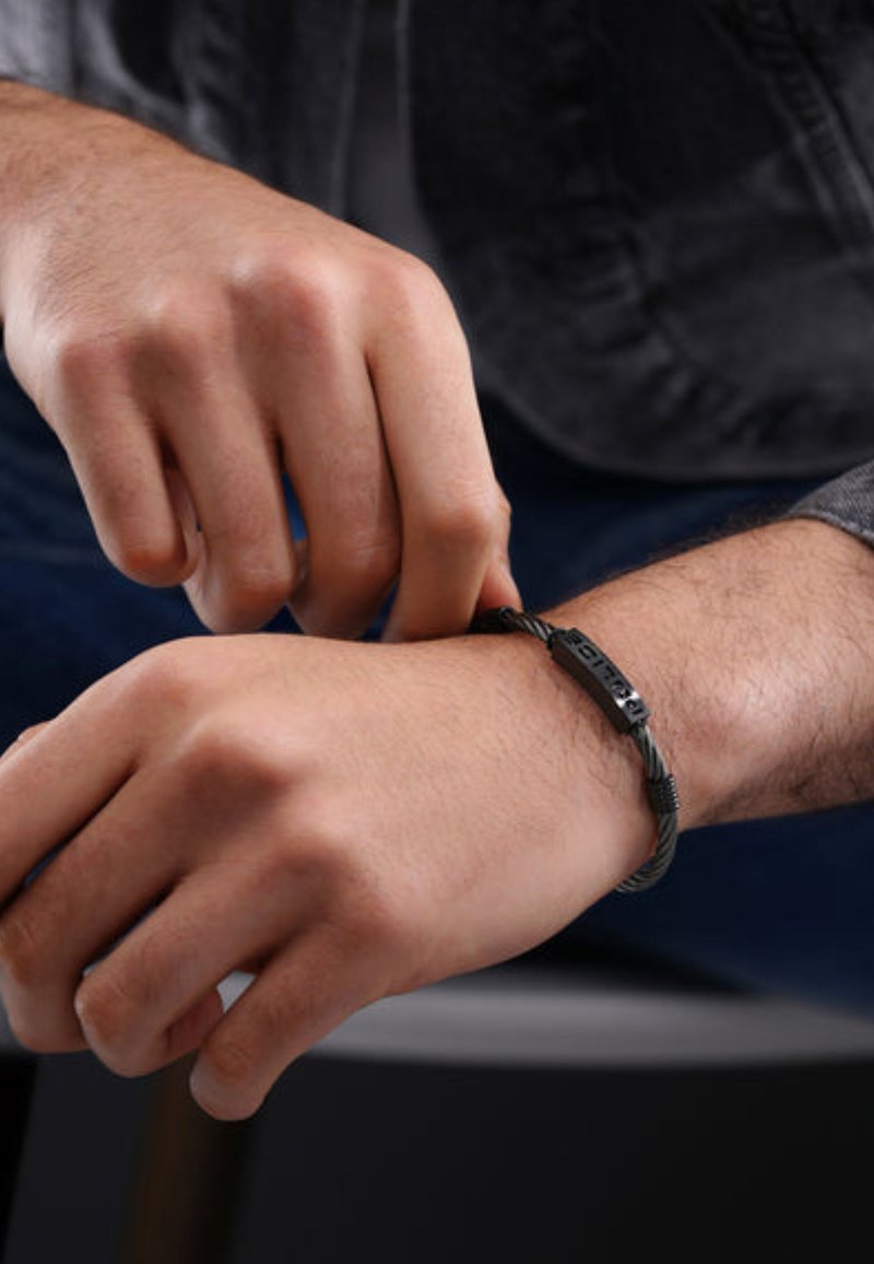 Bracciale intrecciato nero con dettagli testurizzati, dotato di una chiusura piatta. Indossato su un polso, mani che lo regolano. Focus sul design e sul materiale.
