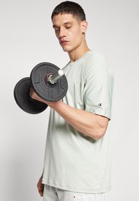 Camiseta de algodón verde claro con mangas cortas, sostenida mientras se levanta un disco de pesas negro con un poste central blanco.