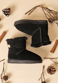 Las botas de tobillo de ante negro presentan un suave forro de piel de lana, una decoración de hardware en forma de copo de nieve y una suela de goma texturizada. Diseñadas con acentos naturales.