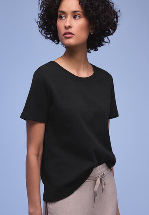 Femme aux cheveux bouclés portant un T-shirt noir uni et un pantalon gris clair à cordon, regardant sur le côté sur un fond bleu clair.