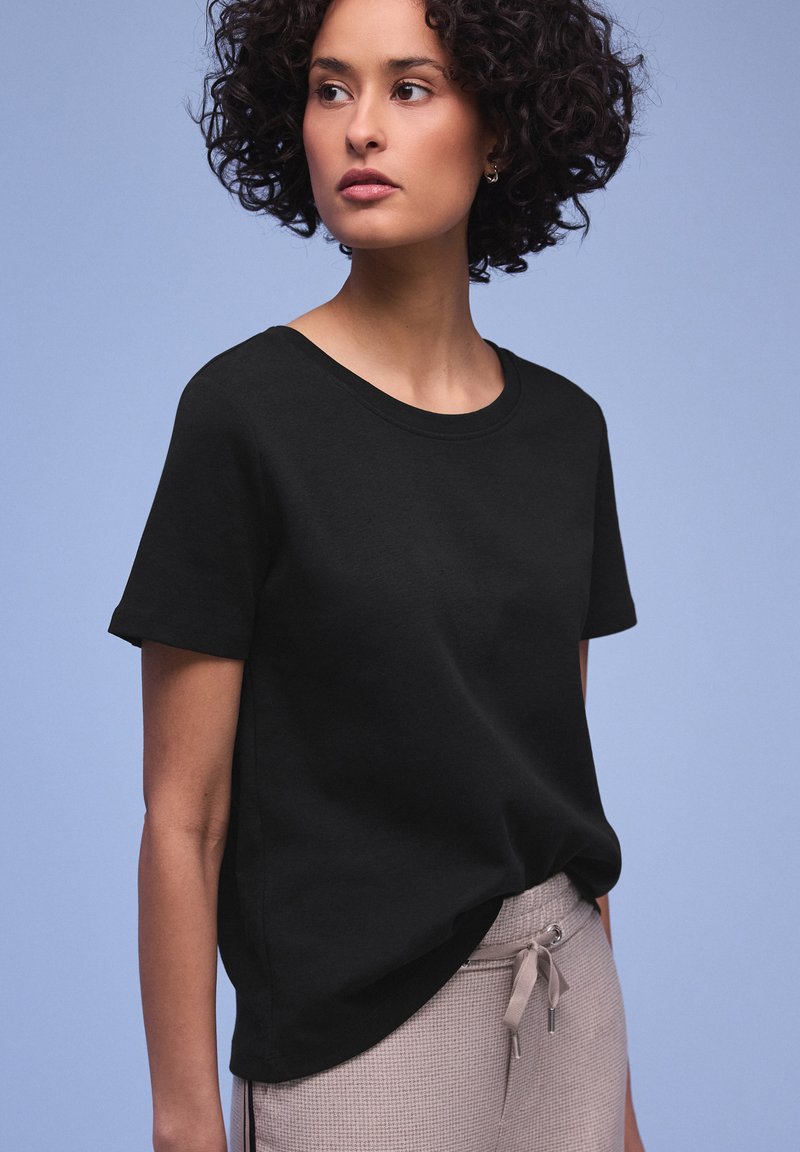 Femme aux cheveux bouclés portant un T-shirt noir uni et un pantalon gris clair à cordon, regardant sur le côté sur un fond bleu clair.