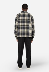 Kockás flanel ing nádfekete és krém színben, melynek dobozos fazonja, gombos mandzsettái és egyenes alja van. Fekete, bő szárú nadrággal párosítva.