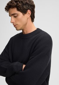 Maglione nero in maglia con un motivo a coste testurizzato, scollo rotondo e maniche ampie, indossato da un modello con le braccia incrociate su uno sfondo neutro.