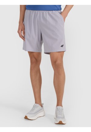 Hombre con pantalones cortos deportivos gris claro con el logo 4F y zapatillas blancas, de pie con una mano en el bolsillo sobre fondo liso.