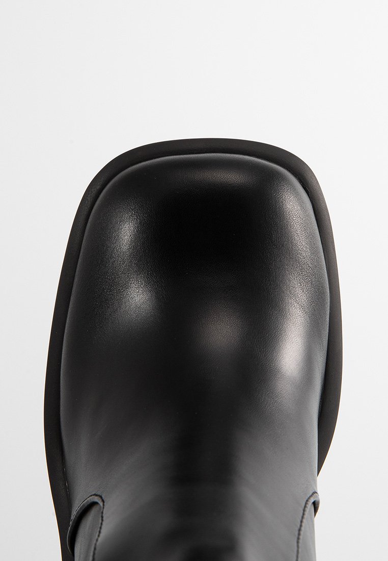 Bottine en cuir noir avec un bout carré et une texture lisse, photographiée de dessus sur un fond clair et uni.