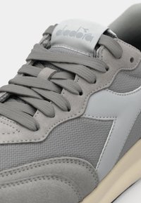 Chaussure de sport grise présentant une combinaison de matériaux en mesh et en daim ; inclut des lacets ton sur ton et une semelle en caoutchouc. Logo affiché sur la languette.
