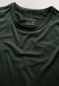 T-shirt de sport vert foncé avec col rond indiquant la taille moyenne et étiquette de marque "Next Active" imprimée à l'intérieur du col.