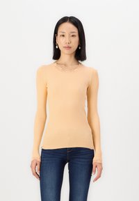 Geribbelde langemouwen top in licht perzik met een geschulpte halslijn en textiele stof. Gecombineerd met strakke donkere jeans.