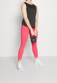 Czarny top bez rękawów, jasnoróżowe legginsy, białe buty sportowe. Trzyma czarny kettlebel z niebieskim akcentem. Gładkie tekstury, dopasowany krój.