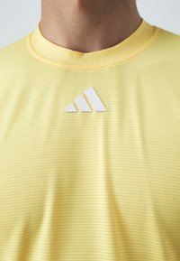 Camiseta atlética amarilla con textura acanalada, cuello redondo y logo blanco de adidas en el pecho. Tejido suave y diseño ajustado.