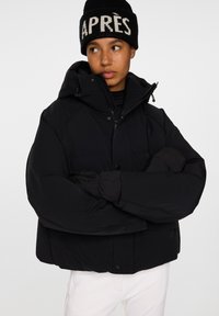 Svart puffjacka med huva och tryckknappar, med elastiska ärmslut. Bärs tillsammans med en svart stickad mössa som visar "APRÈS" i vitt.