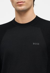 Gros plan d'une personne portant un pull col rond noir avec des épaules texturées et le mot « BOSS » sur la poitrine.