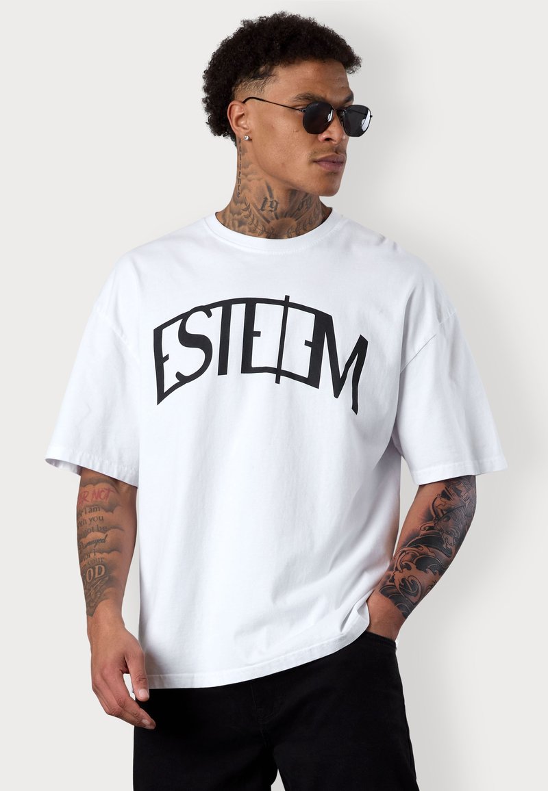 Weißes Baumwoll-T-Shirt mit schwarzem "ESTEEM"-Grafik, lockerer Schnitt, Rundhalsausschnitt, kurze Ärmel, kombiniert mit schwarzen Hosen und Sonnenbrille.