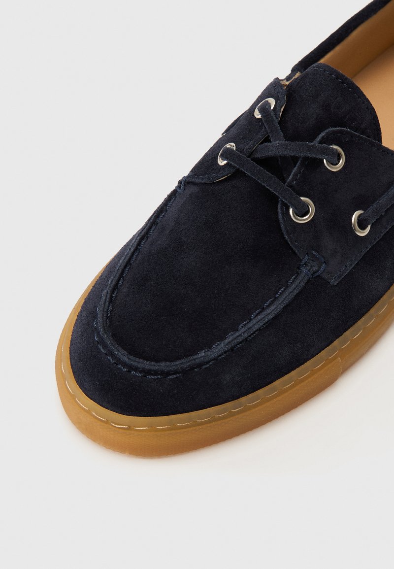 Marineblaue Wildleder-Bootsschuhe mit brauner Gummisohle, versehen mit silbernen Ösen und passenden Schnürsenkeln. Steppnähte entlang der Nähte.