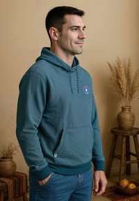 Teal gestreepte hoodie met een voorzak, trekkoord capuchon en geborduurd embleem. Gekocht bij blauwe jeans tegen een neutrale achtergrond.