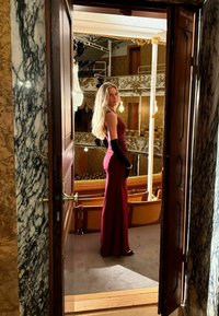 Bordowa suknia wieczorowa na cienkich ramiączkach, gładka w dotyku, uzupełniona czarnymi rękawiczkami, stojąca w drzwiach teatru z ozdobnym wystrojem.