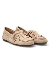 Mocasines de ante beige con flecos y detalles de lazo, suelas planas, mostrados uno al lado del otro sobre un fondo blanco.