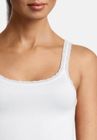 Top bianco senza maniche con bordo di pizzo attorno al collo e alle spalline, realizzato in morbido cotone. Il tessuto appare leggero e aderente.