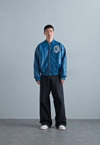 Niebieska skórzana kurtka bombowiec z naszywką astronauty, zapinana na zamek z przodu, w zestawie z czarnymi spodniami o szerokim kroju i białymi sneakersami, stojąca na szarym podłożu.