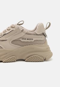 Zapatilla beige con un upper de cuero texturizado y malla, suela de goma gruesa y la marca "STEVE MADDEN" en el lateral.