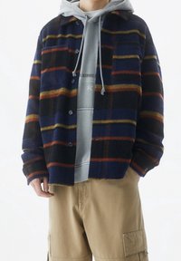 Giacca camicia in pile a quadri nei colori navy, nero e arancione, caratterizzata da bottoni, due tasche frontali e colletto. Indossata sopra un hoodie grigio.
