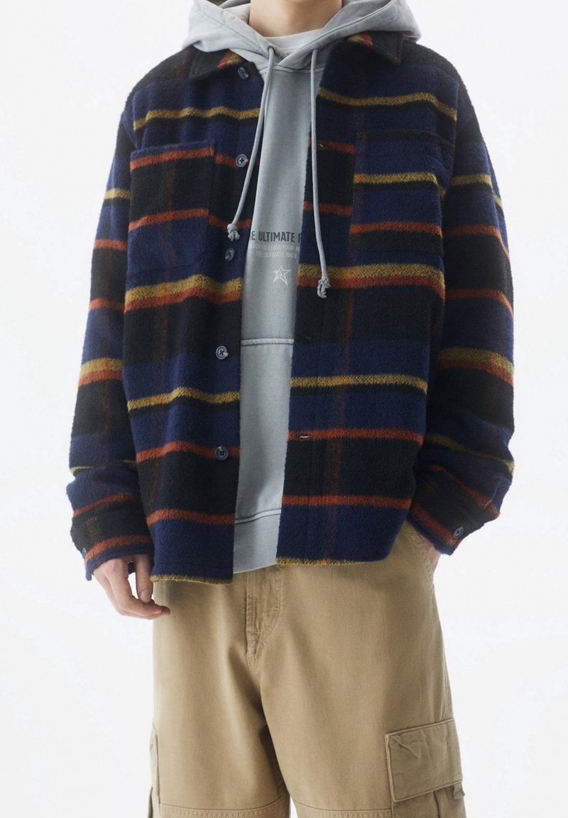 Giacca camicia in pile a quadri nei colori navy, nero e arancione, caratterizzata da bottoni, due tasche frontali e colletto. Indossata sopra un hoodie grigio.